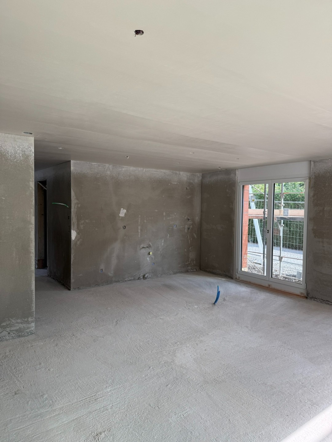 Rohbau eines Wohnzimmers mit unverputzten Wänden und Betonboden, Fenster mit Blick auf den Außenbereich.