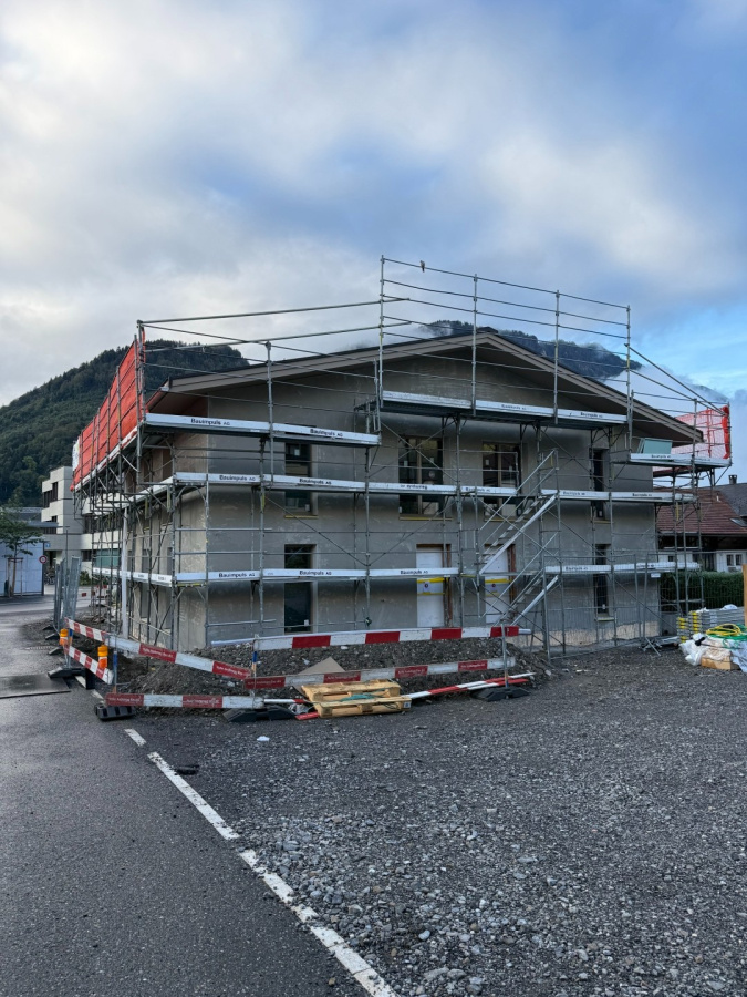 Einfamilienhaus im Bau mit Gerüst, vor Alpenkulisse, Bauphase unter bewölktem Himmel, Baustelle mit Sicherheitsabsperrung.